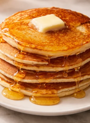 Stack of fluffy homemade pancakes with syrup and fresh fruits on a plate.